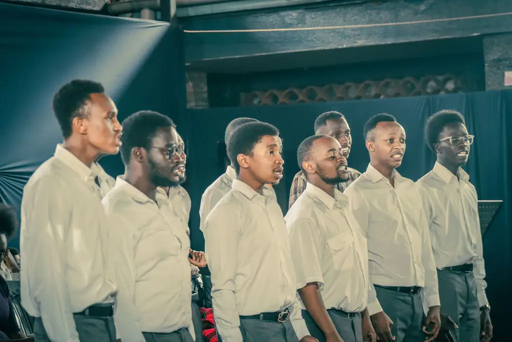 Seven men in white shirts sing side by side, caught mid-note as stage light falls across their faces