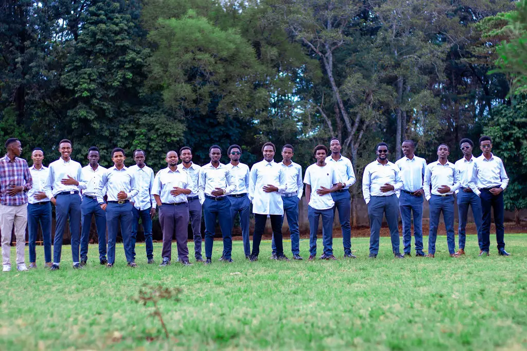 The men of the choir in white shirts and navy trousers, lined up on a green lawn in front of tall trees, hands folded and mid-laugh