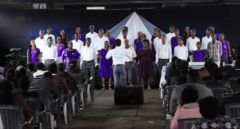 Wide view of an evening concert: the choir in purple and white sings on stage behind a conductor, with the audience seated on rows of chairs under a dark marquee