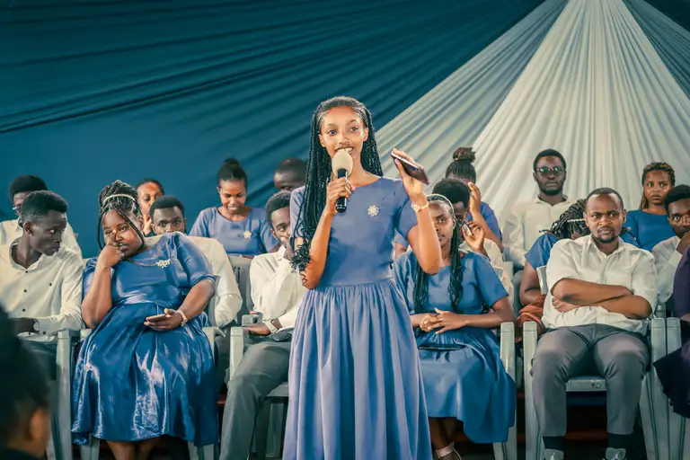 A soloist in a violet dress sings into a microphone, one hand raised, while choir members watch from rows of seats under a navy-and-white draped canopy