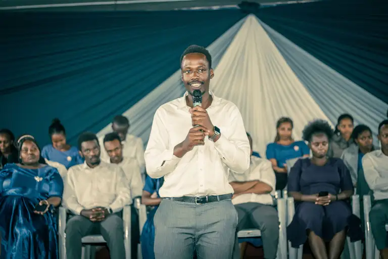 Our chairman stands with a microphone, smiling as he addresses the gathering, with members seated in rows beneath a sunburst of white draping