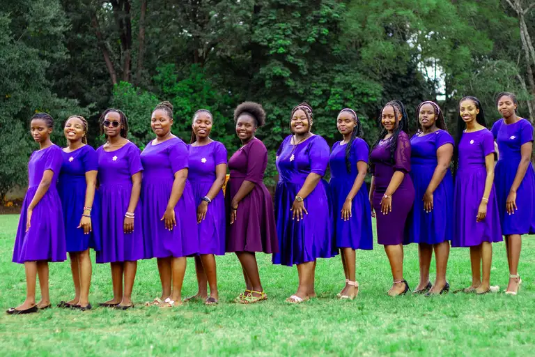 The ladies of the choir in violet and deep-purple dresses, posed in a smiling line on a lawn with lush green trees behind them