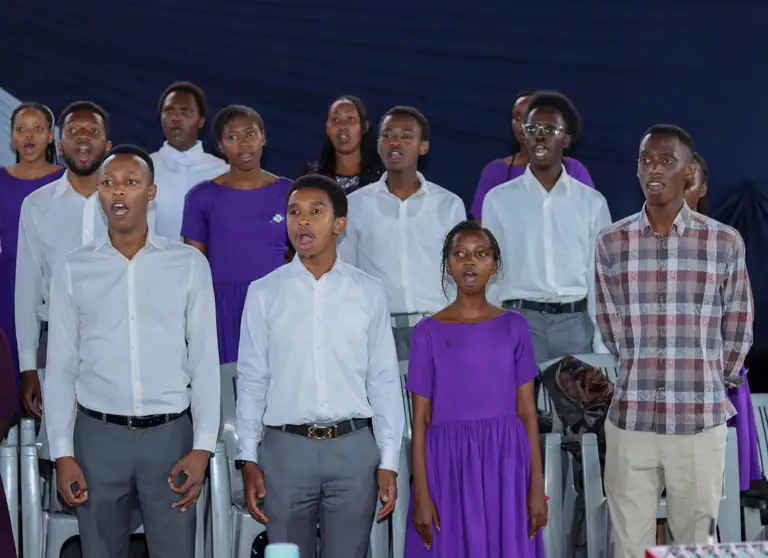 Choir members in white shirts and purple dresses singing mid-phrase in close rows, lit warmly against deep navy draping