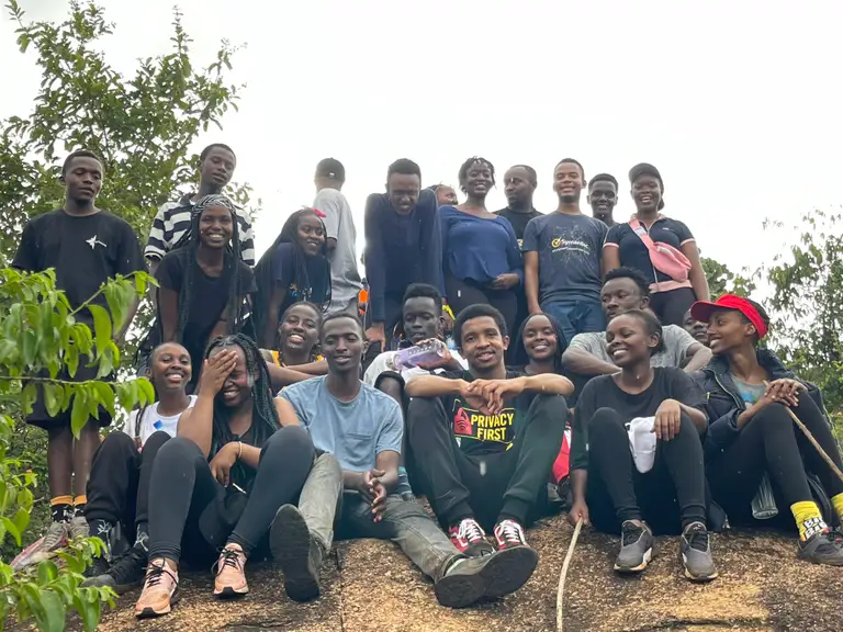 Members in casual hiking clothes crowd together on a rocky outcrop among trees, laughing toward the camera under an overcast sky