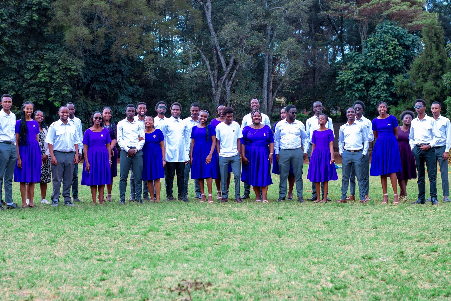 The full choir standing in a long line across a sunlit lawn — women in royal-purple dresses, men in white shirts and grey trousers — with tall trees behind them